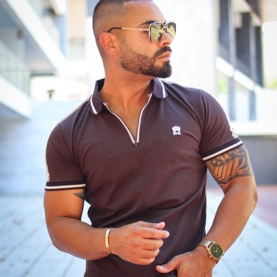 Homem a usar polo castanha com riscas brancas e pretas, em ambiente urbano ao ar livre