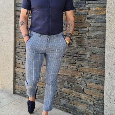 Homem com camisa azul-escura, gravata borboleta e calças xadrez cinza junto parede de pedra.