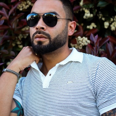 Homem com óculos de sol em armação preta, t-shirt polo branca riscada, pulseira metálica e tatuagens, junto a plantas