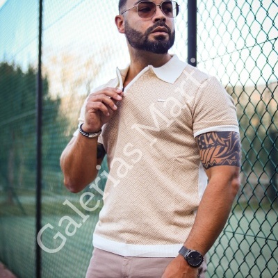 Homem veste polo bege com gola branca, óculos de sol, pulseira e relógio em frente a vedação metálica verde