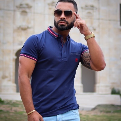 Homem a usar camisa polo azul escura com detalhes vermelhos e brancos e calças azul claro em ambiente exterior