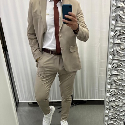 Homem vestido com conjunto formal bege, gravata vermelha e sapatos brancos em frente a um espelho com moldura prateada.