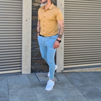 Homem com camisa amarela, calças azuis claras e sapatos brancos encostado a parede de pedra