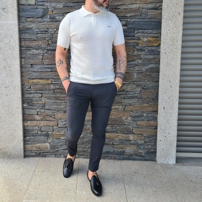 Homem com camisa polo branca e calças pretas junto a parede de pedra cinzenta