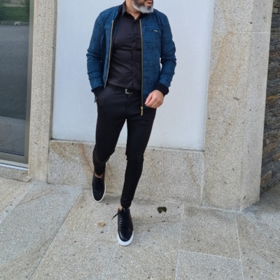Homem com casaco azul, camisa e calças pretas, sapatos pretos com sola branca, em chão de pedra