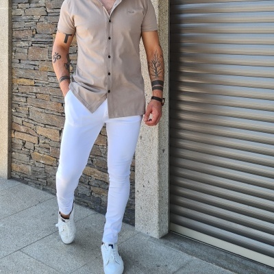 Homem com camisa bege de manga curta e calças brancas junto a parede de pedra e porta metálica