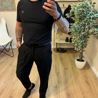 Homem a tirar selfie num espelho com roupa preta, relógio dourado e planta ao fundo