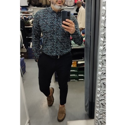 Homem a tirar selfie numa loja de roupa, camisa preta estampada, calças e sapatos castanhos.