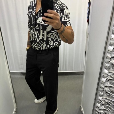 Homem com camisa preta estampada, calças pretas e ténis brancos em frente a espelho