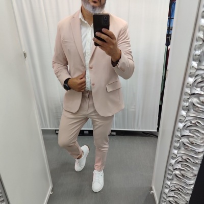 Homem com fato cor de rosa claro, camisa branca e sapatilhas brancas em frente ao espelho.