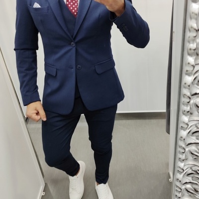 Homem vestido com fato azul escuro, gravata vermelha com bolinhas brancas e sapatos brancos em ambiente interior.