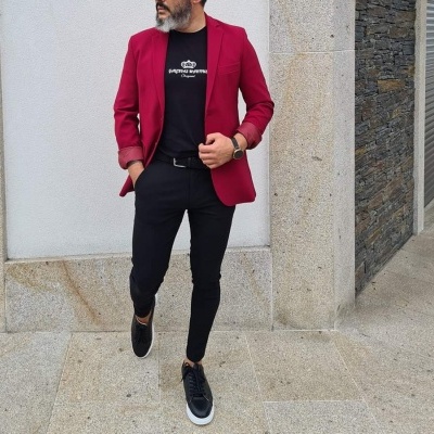 Homem vestido com blusão vermelho, t-shirt preta com texto, calças pretas e ténis pretos junto a parede de pedra cinza