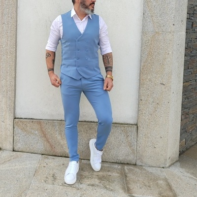 Homem com fato azul claro de colete e calças, camisa branca e sapatos brancos, junto a parede branca e vidro.