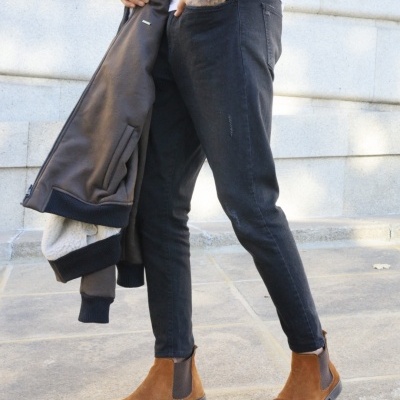 Pessoa com jeans pretos, botas castanhas de camurça e casaco preto segurado em área ao ar livre