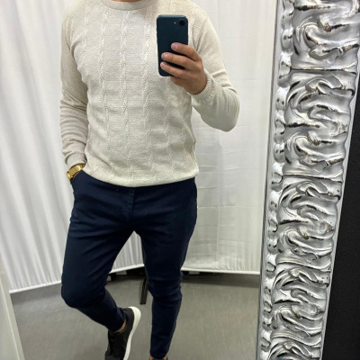 Homem com camisola bege, calças azuis escuras e sapatos pretos tira selfie em espelho prateado.