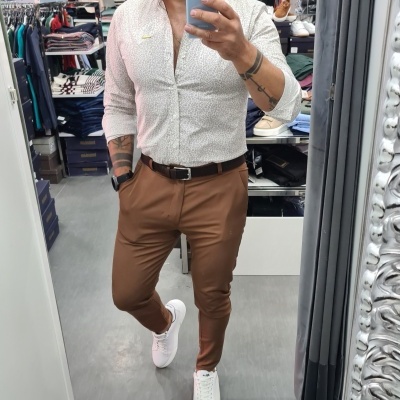 Homem a vestir camisa branca com padrão cinza, calças castanhas, cinto castanho e ténis brancos numa loja de roupa.