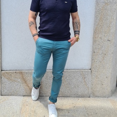 Homem com polo azul escuro, calças azuis claras e sapatilhas brancas contra parede cinzenta