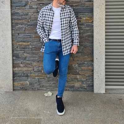 Homem com camisa xadrez, t-shirt branca, jeans azuis, cinto castanho e tênis pretos em frente a parede de pedra