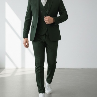 Homem a vestir fato três peças verde escuro, camisa branca e gravata cinza claro com padrão, calças verdes e sapatos brancos numa sala clara minimalista.