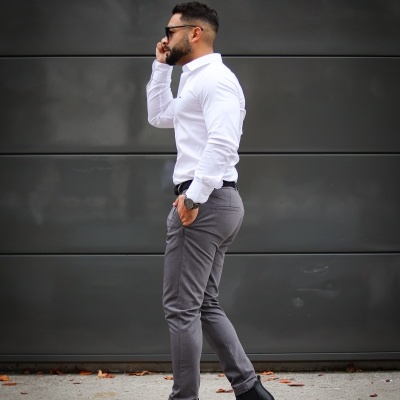 Homem com camisa branca, calças cinzentas e botins pretos falando ao telefone junto a uma parede cinzenta