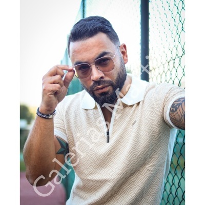 Homem com camisa polo bege clara e óculos de sol ao lado de vedação verde