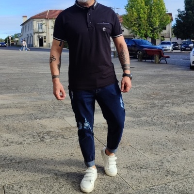 Homem com polo preta, jeans azul e sapatilhas brancas numa rua urbana