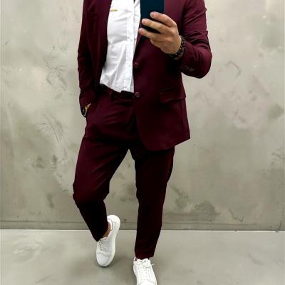 Homem vestido com fato vinho e camisa branca tirando selfie em fundo de parede de cimento.