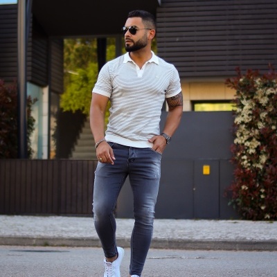 Homem vestido casual com pólo branco, calças de ganga e sapatilhas brancas em área urbana moderna.