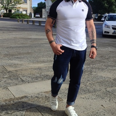 Homem com roupa casual composta por polo branco e preto, jeans com rasgões, e sapatilhas brancas num cenário urbano