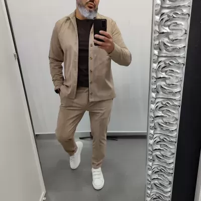 Homem com fato bege, t-shirt castanha e ténis brancos em frente a espelho com moldura prateada