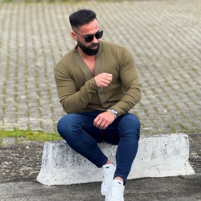 Homem sentado com cardigan verde caqui, calças azuis, ténis brancos e óculos de sol.