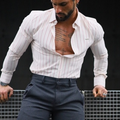 Homem com camisa branca às riscas vermelhas e calças cinza encostado a uma grade metálica