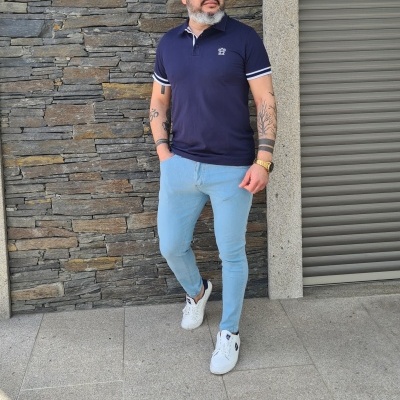 Homem com polo azul escuro e calças azul claro à frente de parede de pedra