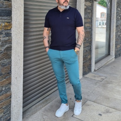 Homem com polo preta e calças azuis claras junto a parede de pedra