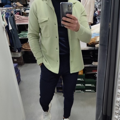 Homem com camisa verde clara, t-shirt preta, calças pretas e sapatilhas brancas a tirar selfie numa loja de roupa