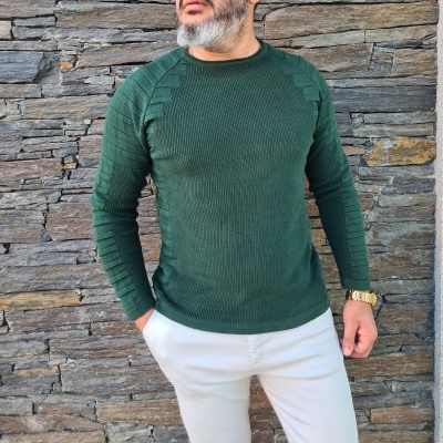 homem a usar camisola verde escura de malha com padrão texturizado e calças brancas frente a parede de pedra cinza