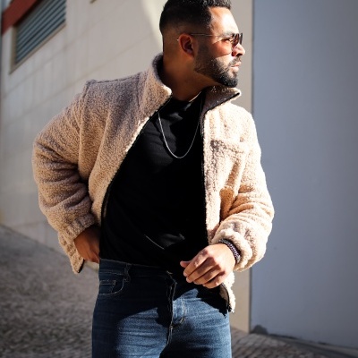 Homem de colete bege de pelo e óculos de sol em ambiente urbano.