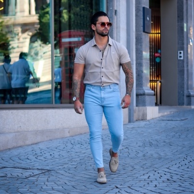 Homem com camisa bege, jeans azul claro e sapatos bege a caminhar numa rua urbana.