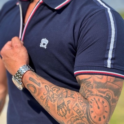 Homem com polo azul escuro com riscas e bordado, braço tatuado e relógio prateado