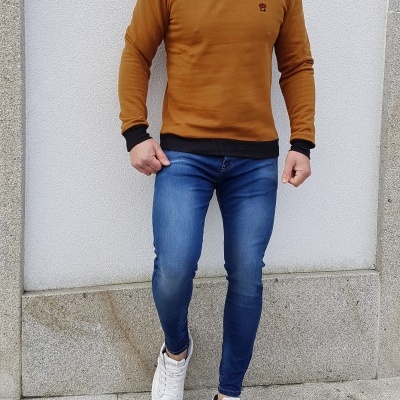 Homem com camisola castanha, calças de ganga azuis e ténis brancos em fundo de parede de pedra.