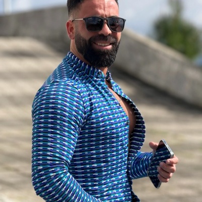 Homem com camisa azul de padrão geométrico e óculos de sol sentado ao ar livre em dia ensolarado.