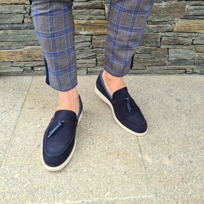 Sapatos tipo mocassim castanhos escuros com tassel e sola branca, em piso de pedra e parede de pedra ao fundo