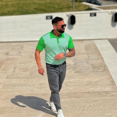 Homem com polo verde e calças cinzentas numa área exterior com carros ao fundo