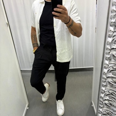 Homem de pé em frente ao espelho com camisa branca, t-shirt preta, calças pretas e ténis brancos.