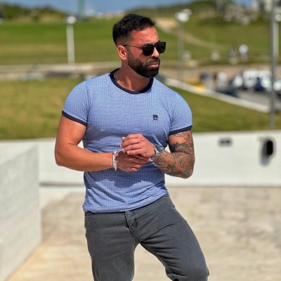 Homem a usar t-shirt azul clara com padrão listrado fino e logo AK no peito, calças cinzentas, óculos de sol e pulseira, ao ar livre