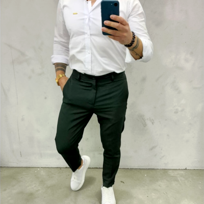 Homem com camisa branca, calças pretas e sapatilhas brancas em fundo cinza a tirar selfie.