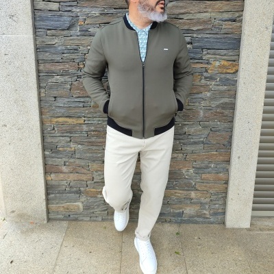 Homem com casaco verde-oliva, calças bege e ténis brancos junto a parede de pedras.