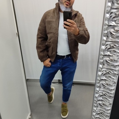 Homem com casaco castanho felpudo, calças azuis e sapatilhas verdes a tirar selfie no espelho