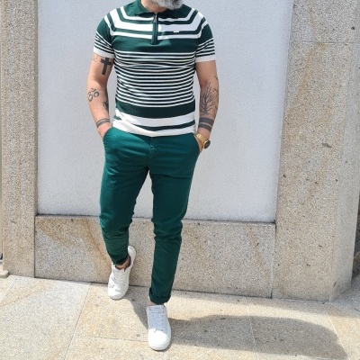 Homem a usar t-shirt polo listrada, calças verdes e sapatilhas brancas junto a uma parede branca e moldura em pedra.