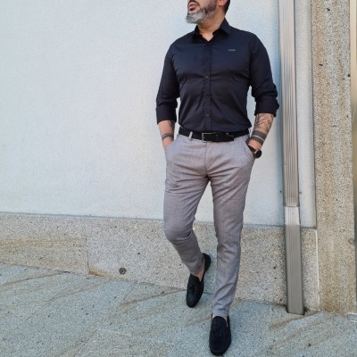 Homem vestido com camisa preta e calças cinza posando contra parede